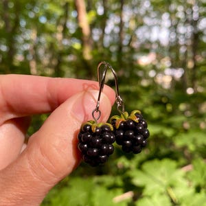 Op de afbeelding: Een paar bungelende oorbellen met realistische, donkerpaarse braambesbedels met kleine groene blaadjes aan de bovenkant. De bessen zijn bevestigd aan zilverkleurige metalen oorhaken, wat een gedetailleerd fruit-thema accessoire vormt.