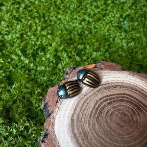 Op de afbeelding: Twee kleine kevervormige ornamenten met iriserende blauwgroene koppen en afgeronde lichamen met afwisselende zwarte en gouden strepen. De kevers zijn geplaatst op een ronde houten schijf met zichtbare jaarringen en ruwe schorsranden.