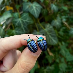 Könnte beinhalten: Ein Paar Ohrstecker in Form von Käfern mit irisierenden blau-grünen Körpern, dunklen vertikalen Streifen und kleinen schwarzen Fühlern. Die Käferfiguren haben eine metallische Oberfläche, die zwischen tiefem Blau und leuchtendem Grün schimmert.