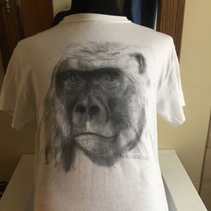 May include: White t-shirt featuring a black and white drawing of a gorilla's face on the front. The drawing is detailed and realistic, with the gorilla's eyes looking directly at the viewer. The text "MARGHERONE" is printed below the drawing.