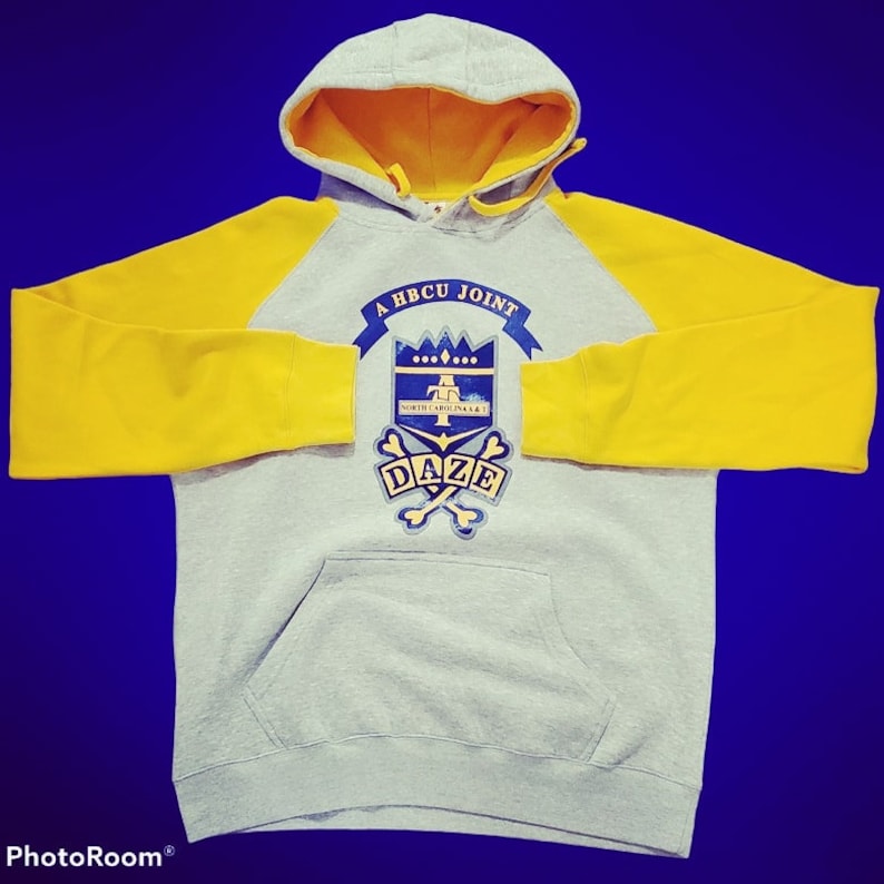Puede incluir: Una sudadera con capucha gris con mangas amarillas. La parte delantera de la sudadera tiene un escudo azul y dorado con el texto "A HBCU JOINT" y "DAZE" en &eacute;l.