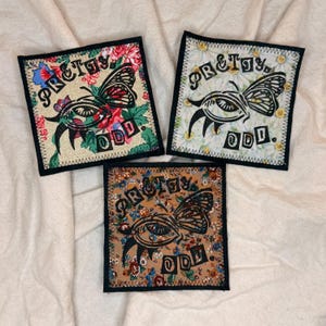 May include: Three square patches with a black border and the words "PRETTY ODD" in black. Each patch features a butterfly design with an eye in the center. The patches have floral backgrounds in various colors.