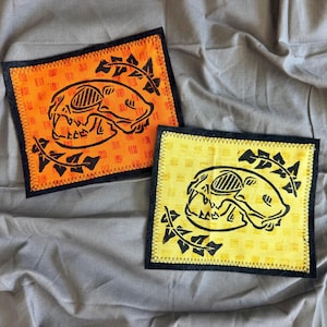 Könnte beinhalten: Zwei quadratische Stoffaufnäher mit schwarzen, genähten Rändern. Ein Aufnäher ist leuchtend orange und der andere gelb, beide mit einem schwarzen Druck im Tintenstil eines Schädels, der von blättrigen Ranken umgeben ist. Der Stoff hat eine feine Gitterstruktur.
