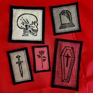 May include: Five rectangular fabric patches with black borders and stitched edges. The designs include a black skull on cream, a tombstone on grey, a dagger on tan, a rose on pink, and a coffin with a cross on red fabric. Each patch features a distinct graphic.