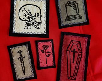 May include: Five rectangular fabric patches with black borders and stitched edges. The designs include a black skull on cream, a tombstone on grey, a dagger on tan, a rose on pink, and a coffin with a cross on red fabric. Each patch features a distinct graphic.