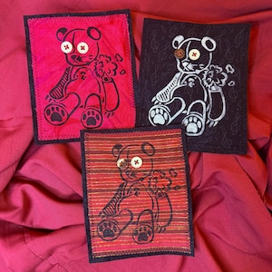 Op de afbeelding: Drie rechthoekige stoffen patches met berenillustraties. Eén patch is felroze, één is zwart met muzieknoten en één heeft rode en gouden horizontale strepen. Elke beer heeft twee knoopogen en een gestikte rand rondom.