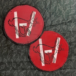 Op de afbeelding: Twee ronde, rode patches met een zwarte rand. Elke patch heeft een wit konijn en een witte verticale lijn met een horizontale lijn erdoor. De patches liggen op een donkere achtergrond met een muzieknotenpatroon.