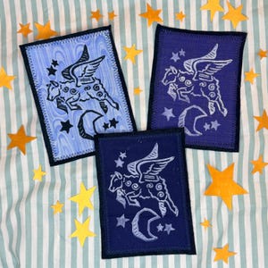 May include: Three rectangular patches with a winged sheep design, stars, and a crescent moon. The patches are in shades of blue and purple, with black borders and white stitching. Scattered around are yellow star-shaped cutouts.