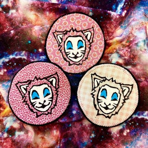May include: Three round fabric patches with a lion face design. Two patches have a pink floral pattern, and one has a pink and white gingham pattern. The lion's face is black and white with blue eyes.