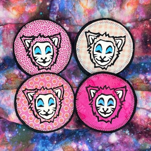 May include: Four circular patches featuring a cartoon lion face. Each patch has a different pink patterned background, including floral, leopard print, and solid pink. The lion's face is white with blue eyes and a black outline.