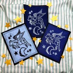 May include: Three rectangular patches with a winged sheep design in light blue and navy blue, with a black border. The patches are surrounded by gold star cutouts, all on a striped background.