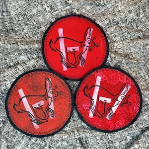 May include: Three red circular patches with a black stitched border. Each patch features a black rabbit illustration with a white vertical stripe and a white horizontal stripe. The patches are on a textured gray surface.
