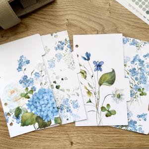 May include: Four white planner inserts with floral designs. The inserts feature watercolor illustrations of blue hydrangeas, roses, and other flowers. The inserts have a tab at the top and are arranged on a wooden surface.