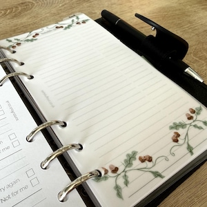 May include: A ring-bound planner insert featuring horizontal lines and a decorative border of brown acorns and green oak leaves. The translucent page includes the text sortsstuffout.co.uk and is held by silver metal rings with a black pen attached.