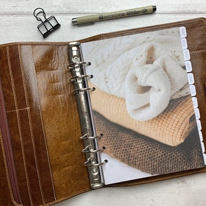 Puede incluir: Carpeta de cuero marrón abierta con mecanismo de anillas plateado. Muestra una página con una foto de jerséis doblados en blanco, beige y marrón. Un clip negro y un bolígrafo sobre una superficie de madera blanca.