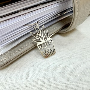 May include: A silver-colored metal charm in the shape of a potted plant, hanging from a ring. The charm is detailed with a plant design and decorative lines. The charm is attached to a planner or notebook.