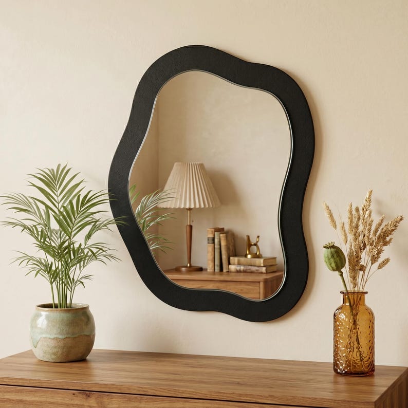 May include: A black, organically shaped wall mirror hangs above a wooden dresser. A potted plant and a vase with dried flowers are on the dresser. The mirror reflects a lamp and books.