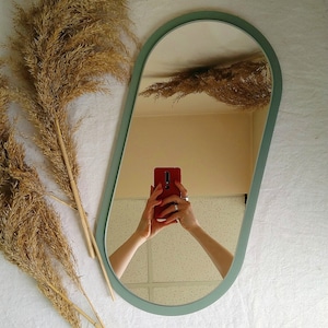 May include: A light green oval mirror with a white frame. The mirror is reflecting a person holding a red phone.