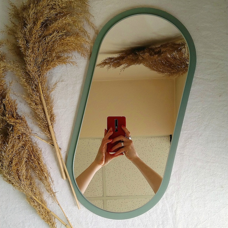 May include: A light green oval mirror with a white frame. The mirror is reflecting a person holding a red phone.