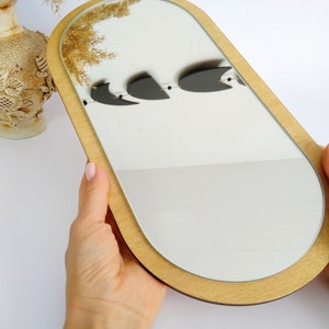 May include: A wooden oval-shaped mirror with a light brown frame. The mirror is held in a person's hands.