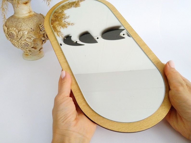 May include: A wooden oval-shaped mirror with a light brown frame. The mirror is held in a person's hands.