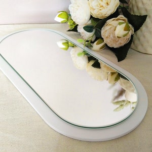 May include: White oval-shaped tray with a mirror inset. The mirror is surrounded by a white frame. The tray is sitting on a white surface.