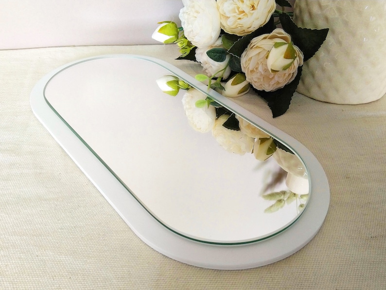 May include: White oval-shaped tray with a mirror inset. The mirror is surrounded by a white frame. The tray is sitting on a white surface.