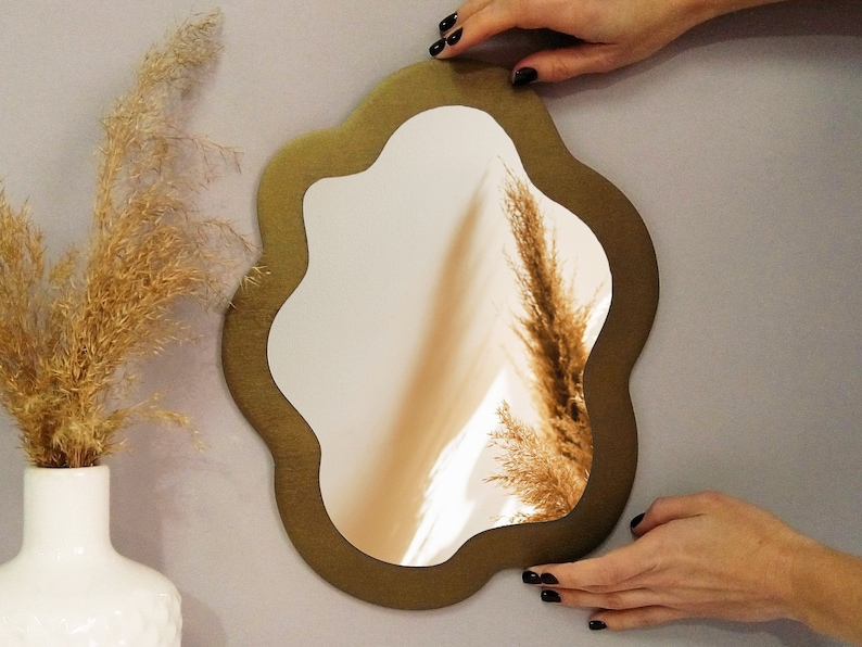 May include: A gold-colored, irregular-shaped mirror with a wavy, organic frame. The mirror is hanging on a light gray wall.