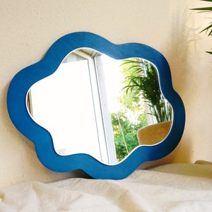 May include: A cloud-shaped mirror with a bright blue frame. The mirror reflects an outdoor scene. The mirror is placed on a white surface. A potted plant is visible in the background. The frame's colour is a vivid blue.