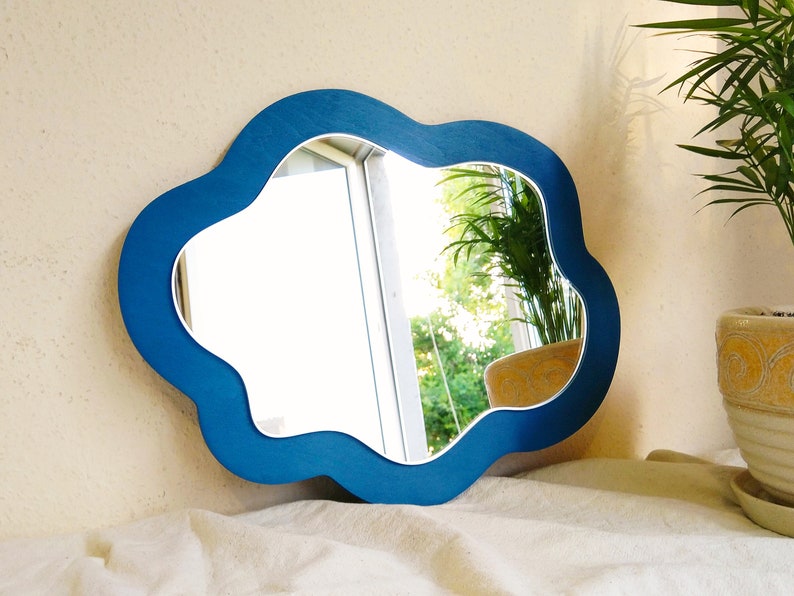 May include: A cloud-shaped mirror with a bright blue frame. The mirror reflects an outdoor scene. The mirror is placed on a white surface. A potted plant is visible in the background. The frame's colour is a vivid blue.
