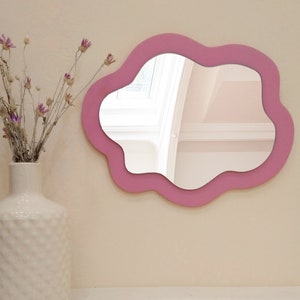 May include: A pink cloud-shaped mirror with a wavy edge hangs on a light beige wall. A white vase with a textured surface holds dried flowers.