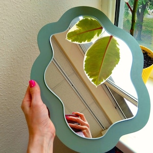 May include: A light blue, wavy-edged mirror with a reflection of a plant with large green leaves. The mirror is being held by a hand with pink nail polish.