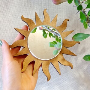 May include: A wooden sun-shaped mirror with a round, reflective surface. The mirror is held in a hand against a light beige background.