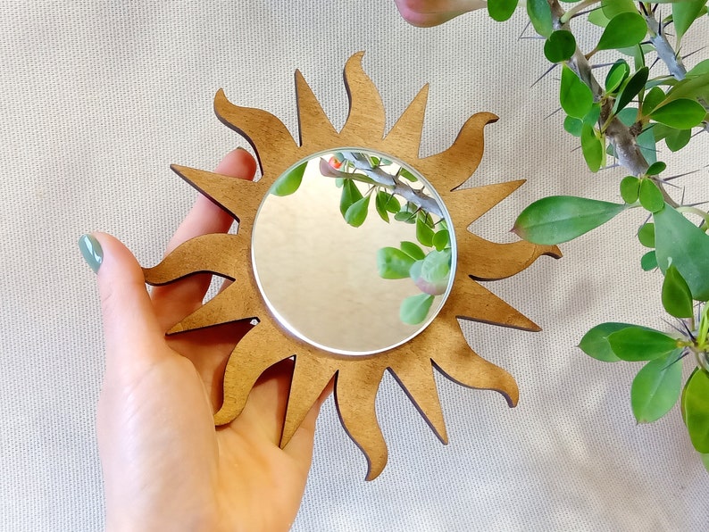 May include: A wooden sun-shaped mirror with a round, reflective surface. The mirror is held in a hand against a light beige background.