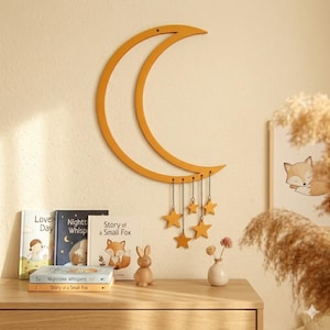 以下が含まれることがあります： 黄色い木製の三日月型の壁掛けで、星の飾りを吊り下げています。三日月はベージュの壁に掛けられており、木製のテーブルの上にあります。「Story of a Small Fox」などのタイトルの本と木製のウサギがテーブルの上にあります。