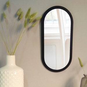 May include: A black oval mirror with a rounded top and bottom. The mirror is hanging on a white wall. There are dried flowers in a white vase to the left of the mirror and dried flowers in a clear glass vase to the right of the mirror.