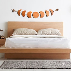 May include: A wooden bed frame with a white duvet and two beige pillows. A wooden moon phase garland hangs above the bed.