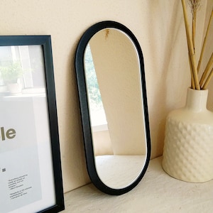 May include: A black oval-shaped mirror with a simple, modern design. The mirror is framed in a thick black wood frame and has a clear reflection.