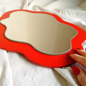 Pu&ograve; includere: Uno specchio rosso di forma irregolare su uno sfondo bianco. Lo specchio ha la forma di una nuvola.