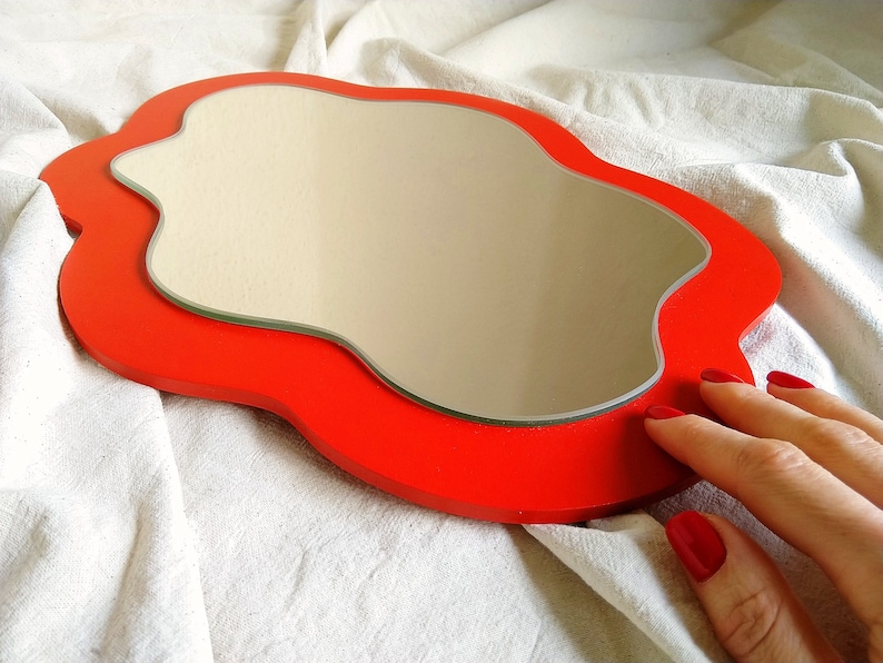 Pu&ograve; includere: Uno specchio rosso di forma irregolare su uno sfondo bianco. Lo specchio ha la forma di una nuvola.