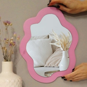 May include: A pink, wavy-shaped wall mirror with a white and beige pillow reflected in the glass.