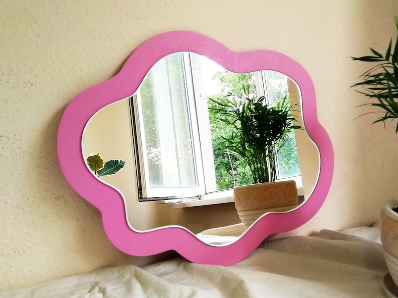 May include: A pink, cloud-shaped mirror with a white inner border. The mirror reflects a window with greenery and a plant in a pot. The mirror is mounted on a cream-coloured wall.