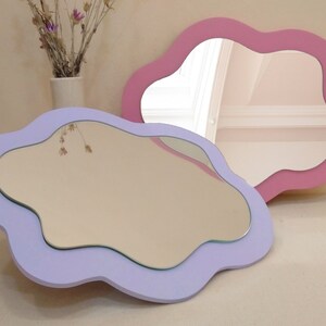May include: Two decorative cloud-shaped mirrors. One is lavender, the other is pink. Both have a smooth, wavy edge and a reflective surface. The mirrors are displayed on a neutral surface, with a vase of dried flowers in the background.