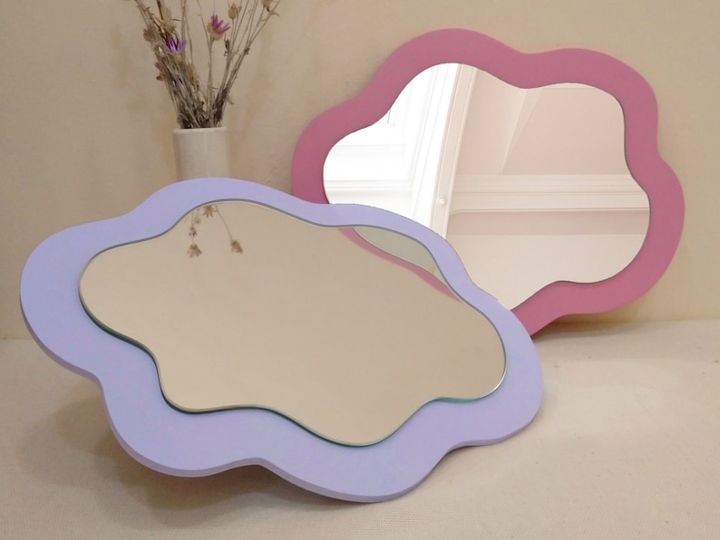 May include: Two decorative cloud-shaped mirrors. One is lavender, the other is pink. Both have a smooth, wavy edge and a reflective surface. The mirrors are displayed on a neutral surface, with a vase of dried flowers in the background.