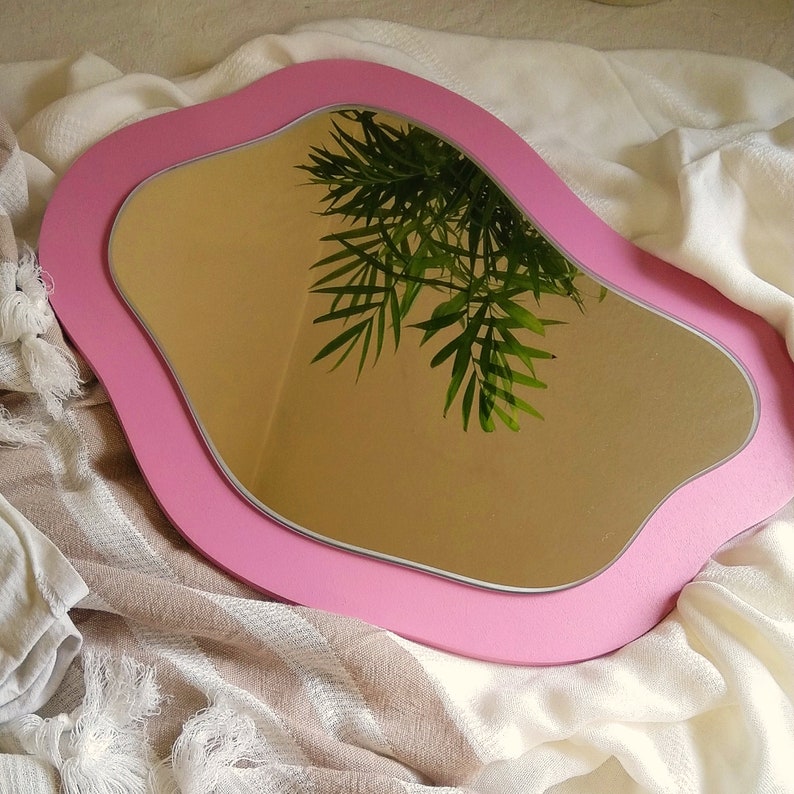 May include: A pink, wavy-edged mirror with a silver trim. The mirror reflects green leafy foliage against a tan wall. The mirror is set on a white and tan striped fabric.