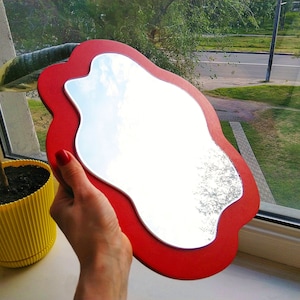 May include: A handheld mirror featuring an irregular, wavy, cloud-like shape with a smooth, bright red frame surrounding a reflective glass surface. The mirror has a distinct, non-symmetrical outline that curves inward and outward along its edges.
