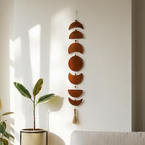 May include: A decorative wall hanging featuring the phases of the moon. The brown metal cutouts are connected by a string and small wooden beads. A tassel hangs from the bottom. The wall hanging is displayed against a white wall.