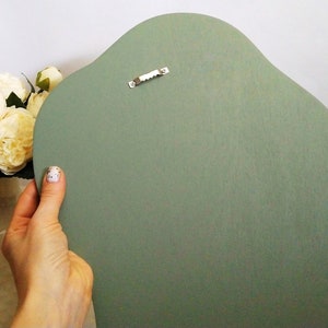 May include: A sage green, irregularly shaped wooden wall hanging. The back of the piece is visible, showing a sawtooth hanger for mounting. The wood has a smooth, matte finish. A hand holds the wall hanging, with a vase of cream-colored flowers in the background.
