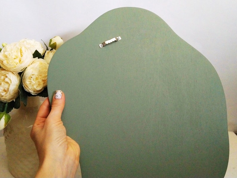May include: A sage green, irregularly shaped wooden wall hanging. The back of the piece is visible, showing a sawtooth hanger for mounting. The wood has a smooth, matte finish. A hand holds the wall hanging, with a vase of cream-colored flowers in the background.