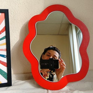 May include: A red, flower-shaped mirror with a wavy edge. The mirror is reflecting a person taking a selfie with a smartphone.